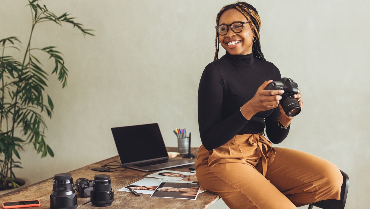 A photographer, perched on the edge of her desk, holds her SLR camera and smiles. Near her are photographs, lenses, and a laptop.