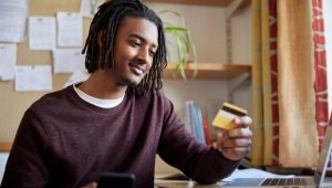 A young man happily contemplates his new credit card with his smartphone in one hand and laptop on the desk in front of him.