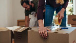 A person seals a cardboard box with packing tape, suggesting they are in the process of moving. The scene includes a man with his hair up adding items to boxes in the background, reinforcing the moving theme. A pair of scissors is visible nearby, likely used for cutting the tape.