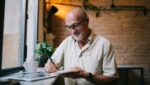 A man sits at a counter in a coffee shop, jotting notes in a spiral-bound notepad; his tablet and a large cup are on the counter in front of him.