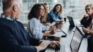 A group of men and women meet at a conference table with laptops and notebooks in front of them.