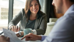 Smiling professional woman in a blazer discussing a document with a colleague across the table in a bright office setting.