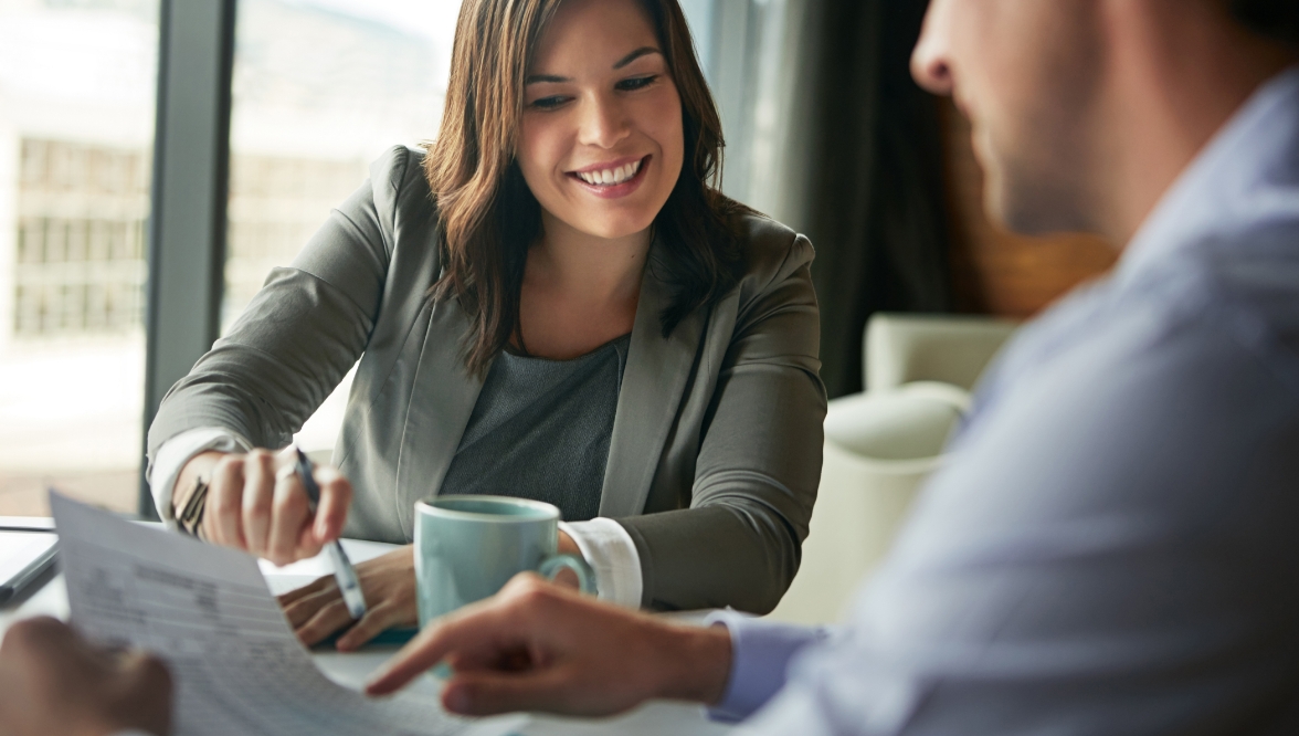 Smiling professional woman in a blazer discussing a document with a colleague across the table in a bright office setting.