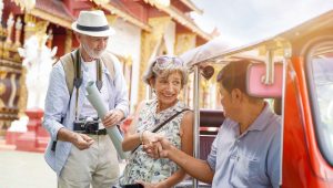 An older couple smiles as the woman shakes hands with a local tuk-tuk driver in front of an ornate temple.