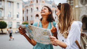 Two young women smiling and looking around while holding a map during their travels in a European city.