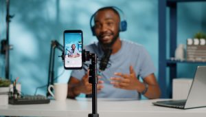 A podcaster, wearing headphones and a light blue T-shirt, gestures to make a point while his image appears on a smartphone in the foreground.