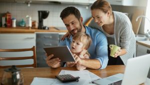 A family of three, a man, a woman, and a toddler, gather around a table with a calculator, papers, and a laptop, suggesting they are managing finances or household matters. The man holds a tablet, while the woman holds a mug.