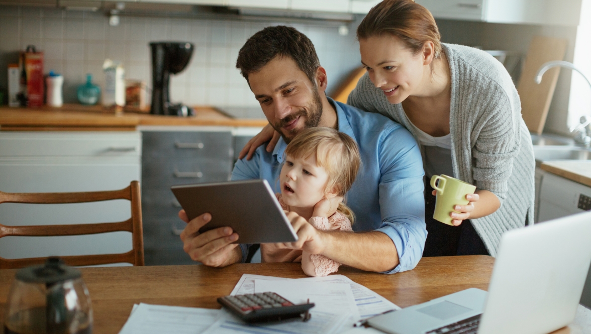A family of three, a man, a woman, and a toddler, gather around a table with a calculator, papers, and a laptop, suggesting they are managing finances or household matters. The man holds a tablet, while the woman holds a mug.