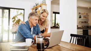 A middle-aged couple looks at a laptop in a kitchen setting. There is a coffee press, a mug, papers, and a book on the wooden table in front of them. The kitchen has white walls and cabinetry, and a doorway leading to another room is visible in the background.