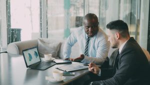 A man in a shirt and tie explains pie charts on a laptop to a bearded man in a suit.