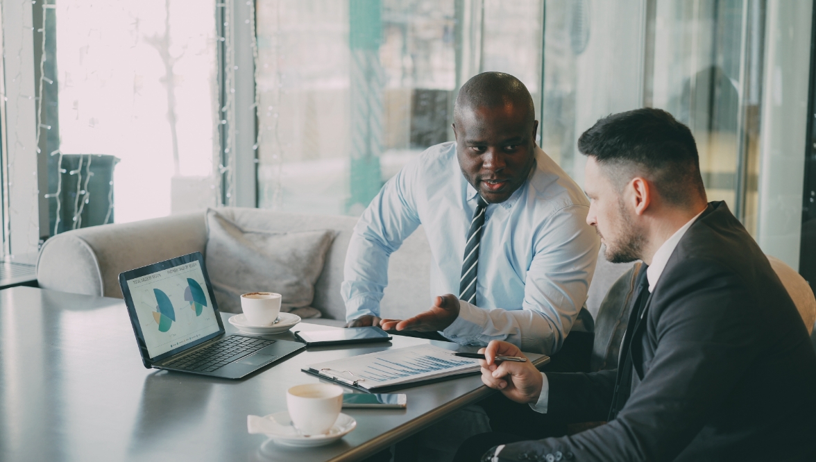 A man in a shirt and tie explains pie charts on a laptop to a bearded man in a suit.
