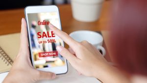 Person holding a smartphone and tapping on an online sale advertisement offering up to 50% off, with a “Shop Now” button displayed on the screen.