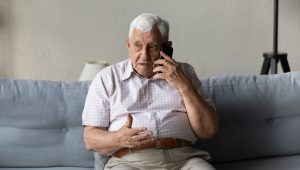 Elderly man with white hair sitting on a couch, holding a mobile phone to his ear with one hand and gesturing with the other, appearing concerned or engaged in conversation.