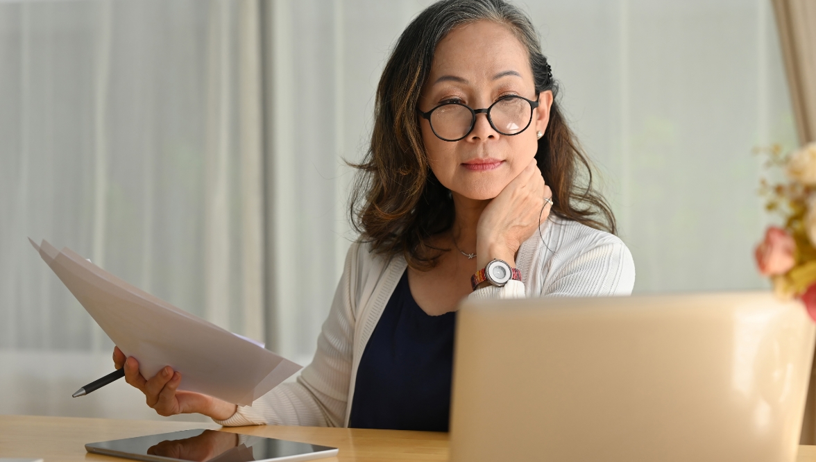A woman with a touch of gray in her hair, wearing glasses and a white cardigan over a navy blue top, looks at a laptop screen. She holds papers and a pen in her right hand and has a tablet on the table in front of her.