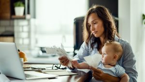 A young mother holding a baby while reviewing receipts and budgeting at home with a laptop.
