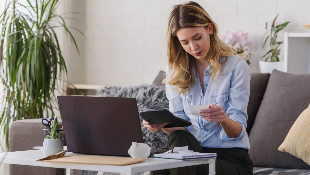 A young woman in a light blue shirt and dark trousers holds a calculator in her right hand and a piece of paper in her left. She is seated on a sofa at a table with a laptop, a manila envelope, a notebook, a plant, and a piggy bank in front of her.