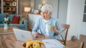 A woman with short, white hair, wearing a light blue cardigan over a white top, holds a mug in one hand and eyeglasses in the other as she consults a laptop. On the table in front of her are papers and a bowl of fruit; a sofa and other living room furniture are in the background.