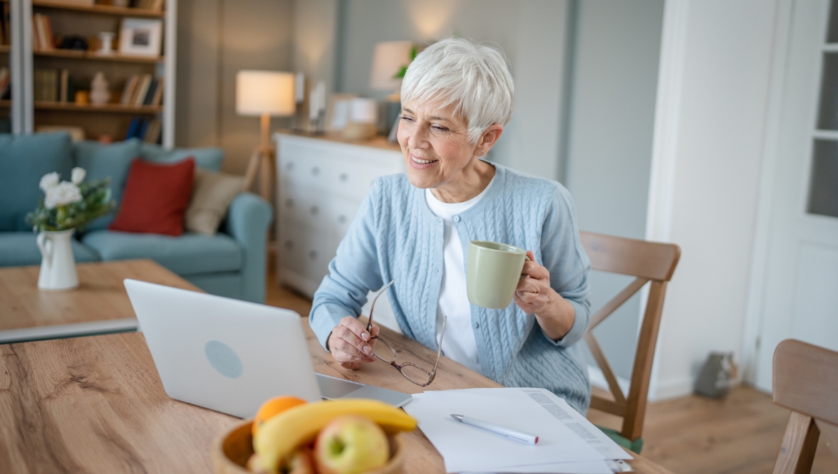 A woman with short, white hair, wearing a light blue cardigan over a white top, holds a mug in one hand and eyeglasses in the other as she consults a laptop. On the table in front of her are papers and a bowl of fruit; a sofa and other living room furniture are in the background.