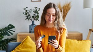 Woman sitting on a couch holding a credit card and smiling while looking at her smartphone, appearing to make an online purchase.