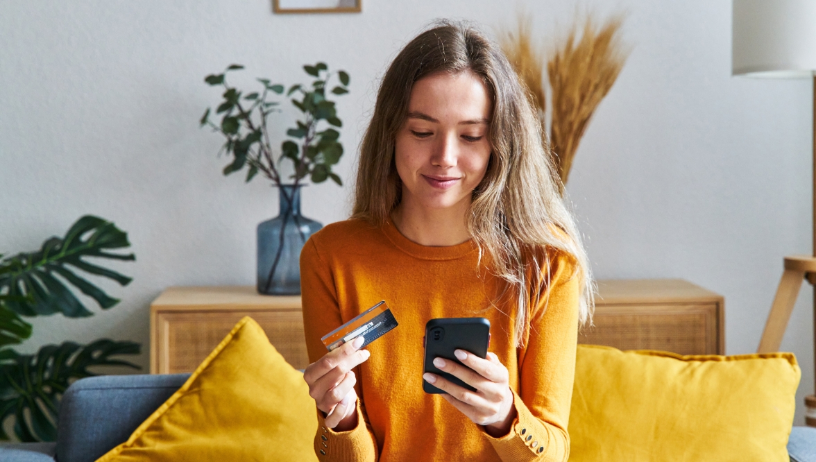 Woman sitting on a couch holding a credit card and smiling while looking at her smartphone, appearing to make an online purchase.