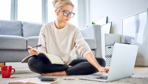 A young blonde woman wearing blue glasses, a cream-colored sweater, and dark leggings sits on the floor surrounded by a laptop, a mug, books, and a calculator. In the background, a gray couch and framed pictures lean against the walls.
