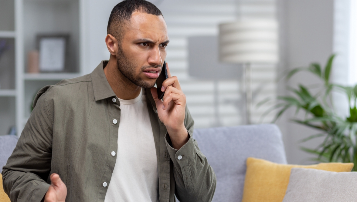 Man sitting on a couch, looking concerned and frustrated while talking on a smartphone.