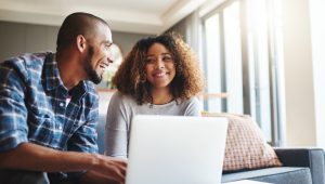 A young man and woman, seated on a gray sofa, smile at each other in a living room while referring to a laptop.