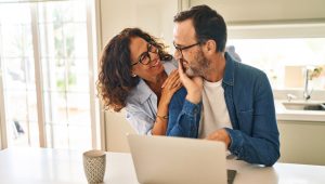 A smiling middle-aged couple wearing glasses share a warm moment in a bright kitchen while sitting at a table with a laptop and coffee mug.