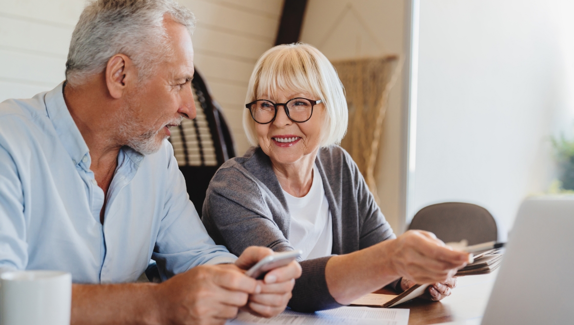 Smiling older couple using a smartphone and laptop together at a table.