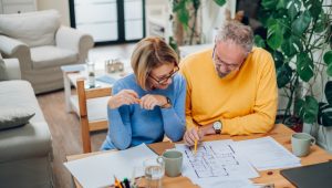 A woman in a blue sweater and a man in a yellow sweater look over a floor plan at a table in a living room setting. Other papers, mugs, a glass, a pen holder, keys, and a laptop are also on the table.