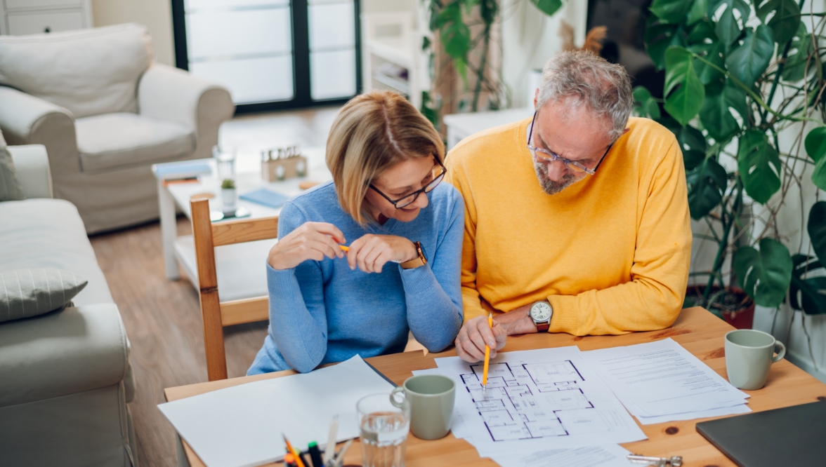 A woman in a blue sweater and a man in a yellow sweater look over a floor plan at a table in a living room setting. Other papers, mugs, a glass, a pen holder, keys, and a laptop are also on the table.