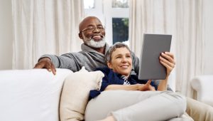 A smiling couple cuddles on a beige sofa; the man looks over the woman’s shoulder at a tablet. Behind them are white-curtained windows.