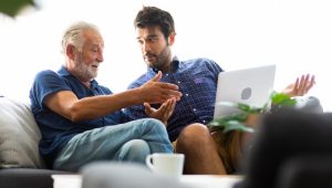 Two men, one older with gray hair and beard and one younger with dark hair and beard, converse on a sofa. The younger man has a laptop on his lap. Each one gestures to the other.