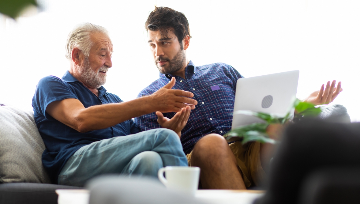 Two men, one older with gray hair and beard and one younger with dark hair and beard, converse on a sofa. The younger man has a laptop on his lap. Each one gestures to the other.