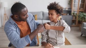 A man in a light blue shirt over a terra cotta-colored T-shirt fist-bumps a child in a khaki shirt. They sit at a counter or a tabletop in a living room setting.