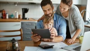 A young woman and a young man with a child on his lap look at a tablet and smile in a kitchen setting. An open laptop, papers, a calculator, and a coffeepot are on the table in front of them.