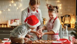 A woman and two children decorate cookies in a kitchen trimmed with holiday lights. The woman and the girl wear matching aprons. The girl also wears a headband with reindeer antlers. The toddler wears a white apron and a Santa hat.