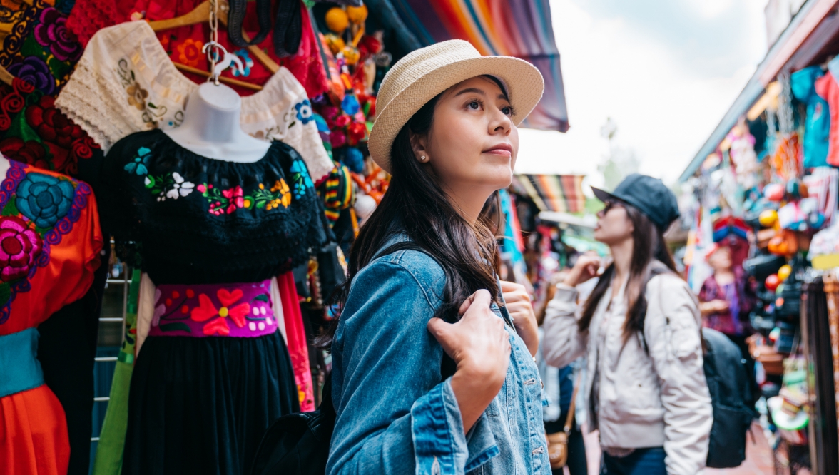 A woman in a hat, wearing a denim jacket and a backpack, looks at merchandise in a colorful bazaar. Behind her, another woman in a baseball cap appears to be shopping.