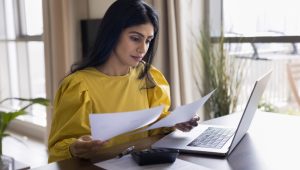 A woman in a yellow top reviews papers at a laptop.