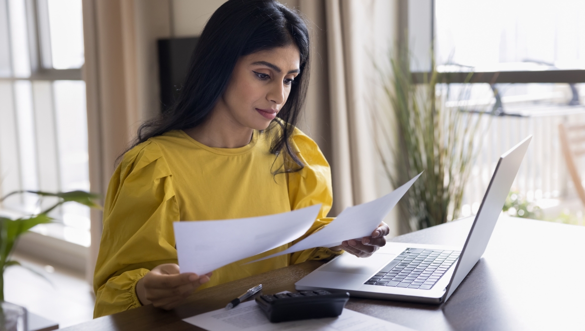 A woman in a yellow top reviews papers at a laptop.