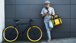 Bicycle courier checking phone while leaning against a wall with delivery bag.