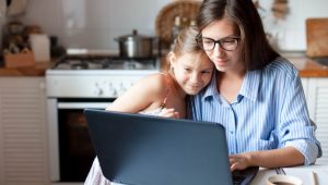 A girl in a sundress snuggles with a woman in glasses and a striped shirt as they look at the screen of a laptop in a kitchen. In the background are counters and a range with a pot on the stove.
