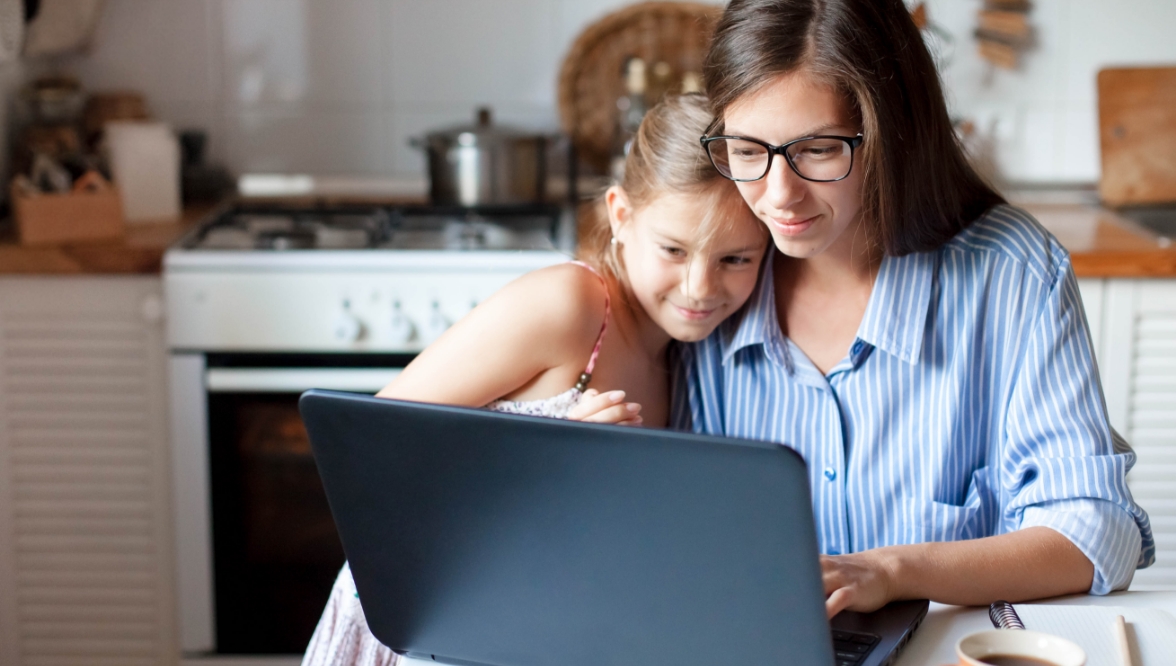 A girl in a sundress snuggles with a woman in glasses and a striped shirt as they look at the screen of a laptop in a kitchen. In the background are counters and a range with a pot on the stove.