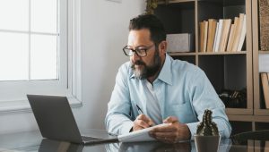 A bearded man in a blue shirt looks at a laptop and writes on paper with a pen. In the foreground is a cactus; in the background are bookshelves.