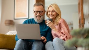 A bearded man with a laptop on his knees and a blonde woman in a pink shirt sit in a living room setting.