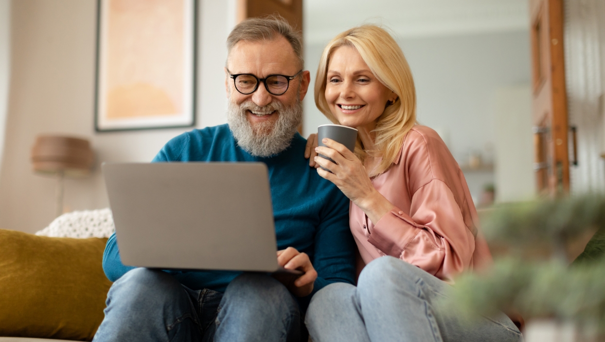 A bearded man with a laptop on his knees and a blonde woman in a pink shirt sit in a living room setting.