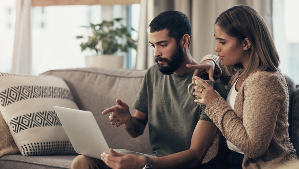 Couple sitting on a couch looking at a laptop together, discussing something with focus.