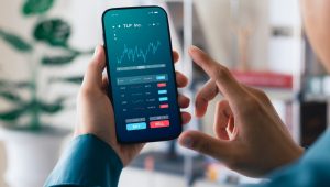 An individual uses a smartphone to monitor stock market trends and trading activity, with a financial app displaying graphs, stock tickers, and options to buy or sell shares.