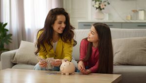 A smiling woman in a yellow cardigan holds a stack of hundred-dollar bills while sitting on a couch next to a young girl in a pink shirt. They look at each other warmly, with a pink piggy bank and more money on the coffee table in front of them, suggesting a conversation about saving money. The background shows a cozy, modern living room with light-colored furnishings and a vase of flowers on a counter.