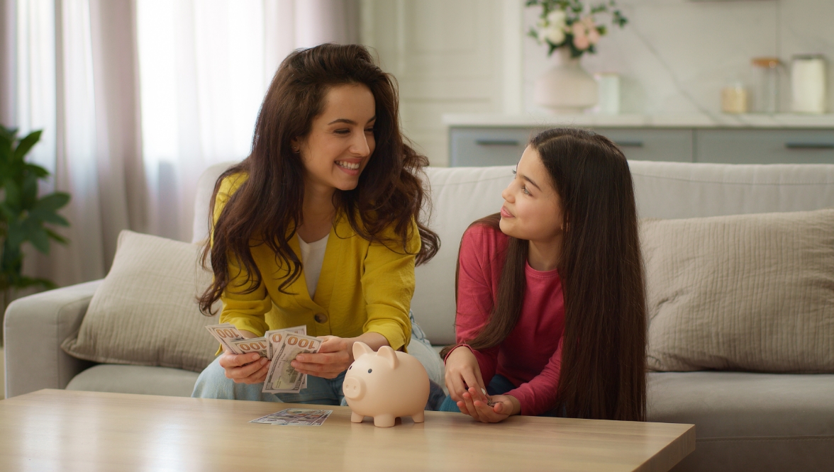A smiling woman in a yellow cardigan holds a stack of hundred-dollar bills while sitting on a couch next to a young girl in a pink shirt. They look at each other warmly, with a pink piggy bank and more money on the coffee table in front of them, suggesting a conversation about saving money. The background shows a cozy, modern living room with light-colored furnishings and a vase of flowers on a counter.